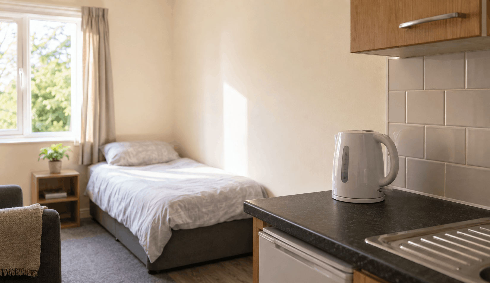 A single room with a made bed by a sunlit window, and a kettle on a kitchen worktop alongside it.